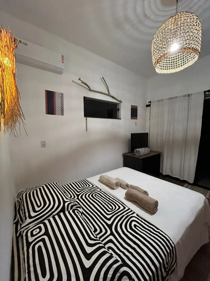 Suíte Alecrim com cama de casal, colcha de padronagem geométrica em preto e branco, televisão na parede e luminária de palha