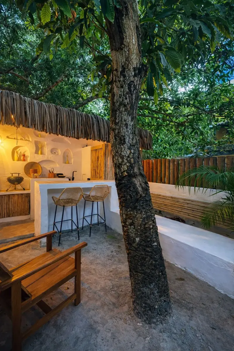 Pátio ao entardecer, com árvore atravessando o deck de madeira e banco comprido junto à cerca de bambu