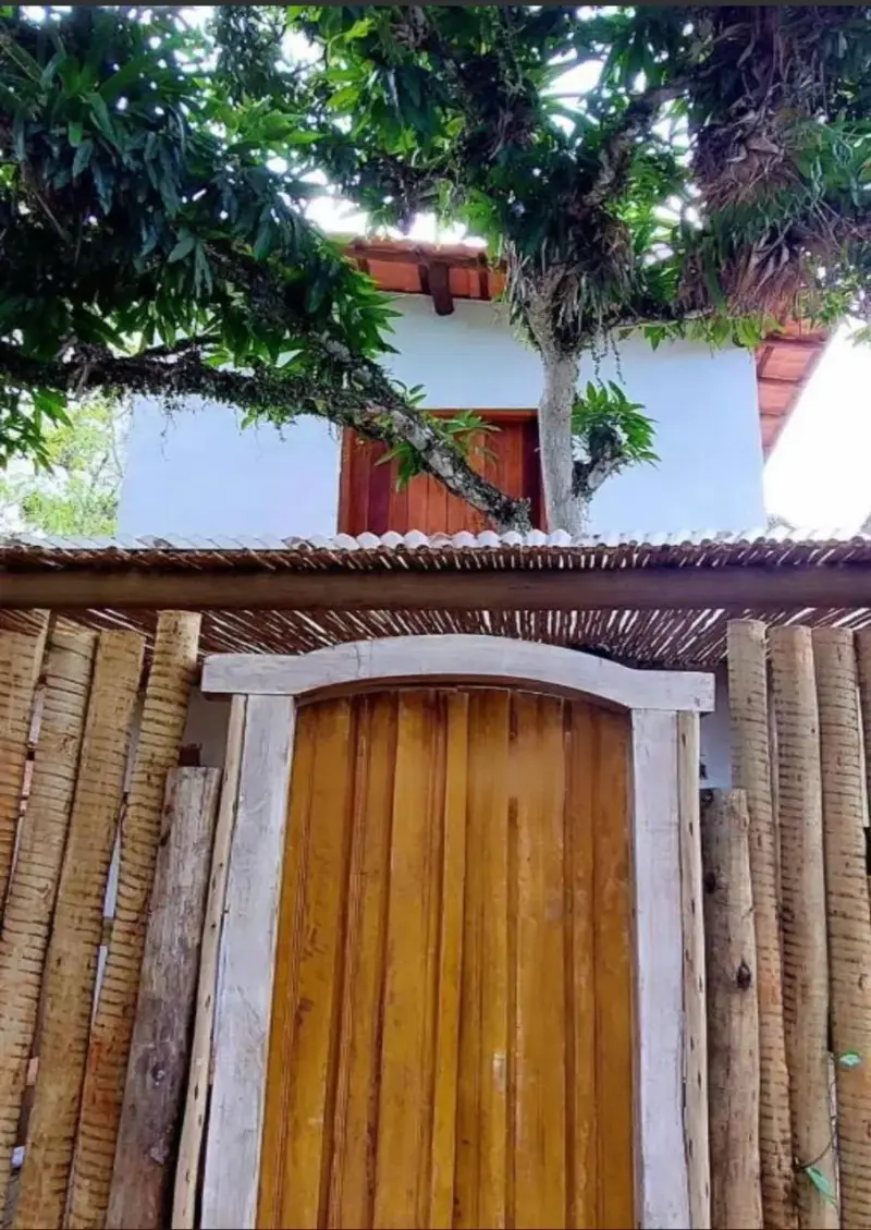 Portão de madeira maciça da Cacimbah entre cercas de bambu, sob uma árvore florida, na rua de areia da vila de Caraíva